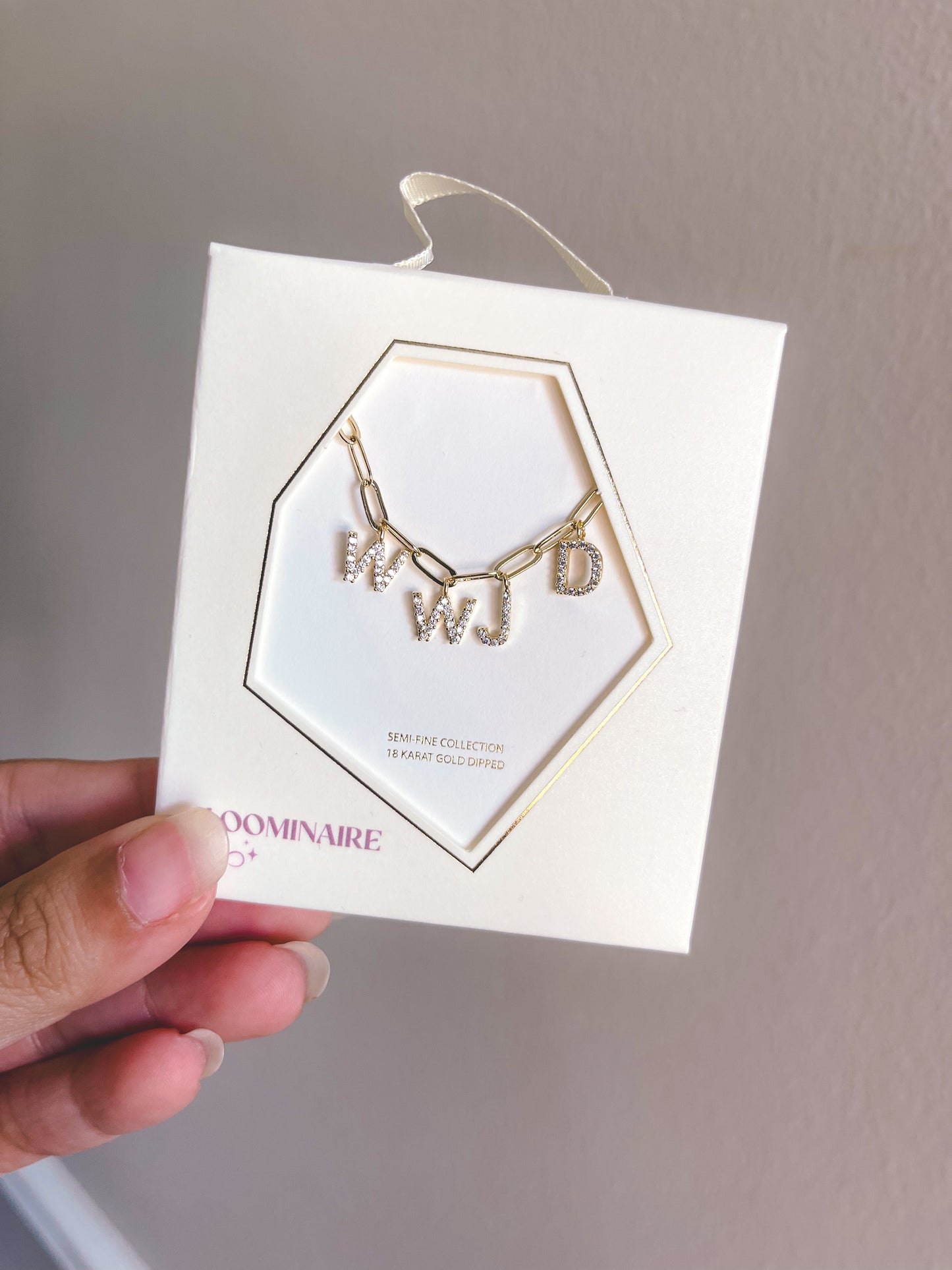 Gold necklace with letter charms on a white card held by a hand against a neutral background