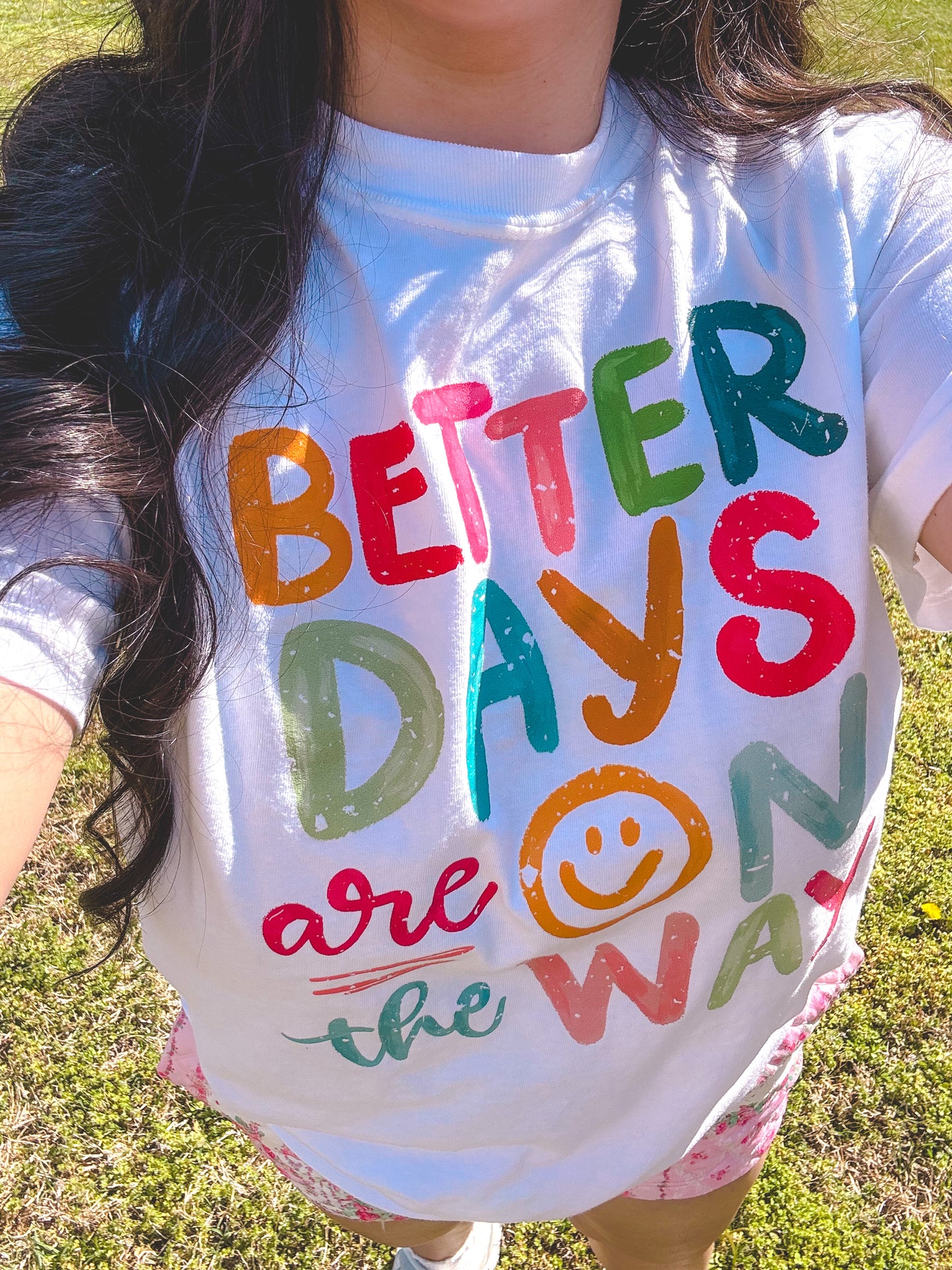 Person wearing a shirt with 'Better Days are on the Way' text in colorful letters outdoors.