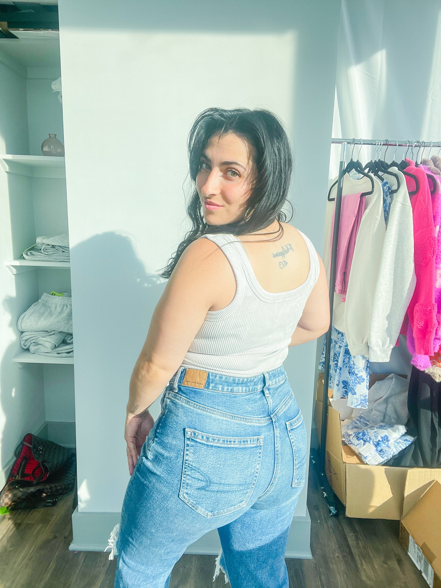 woman standing backwards looking at camera wearing light grey tank top paired with denim jeans