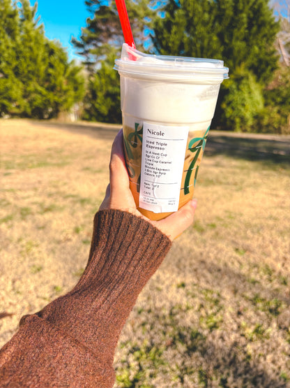 Hand holding a Starbucks iced coffee with a straw outdoors showing brown sleeve on sweater