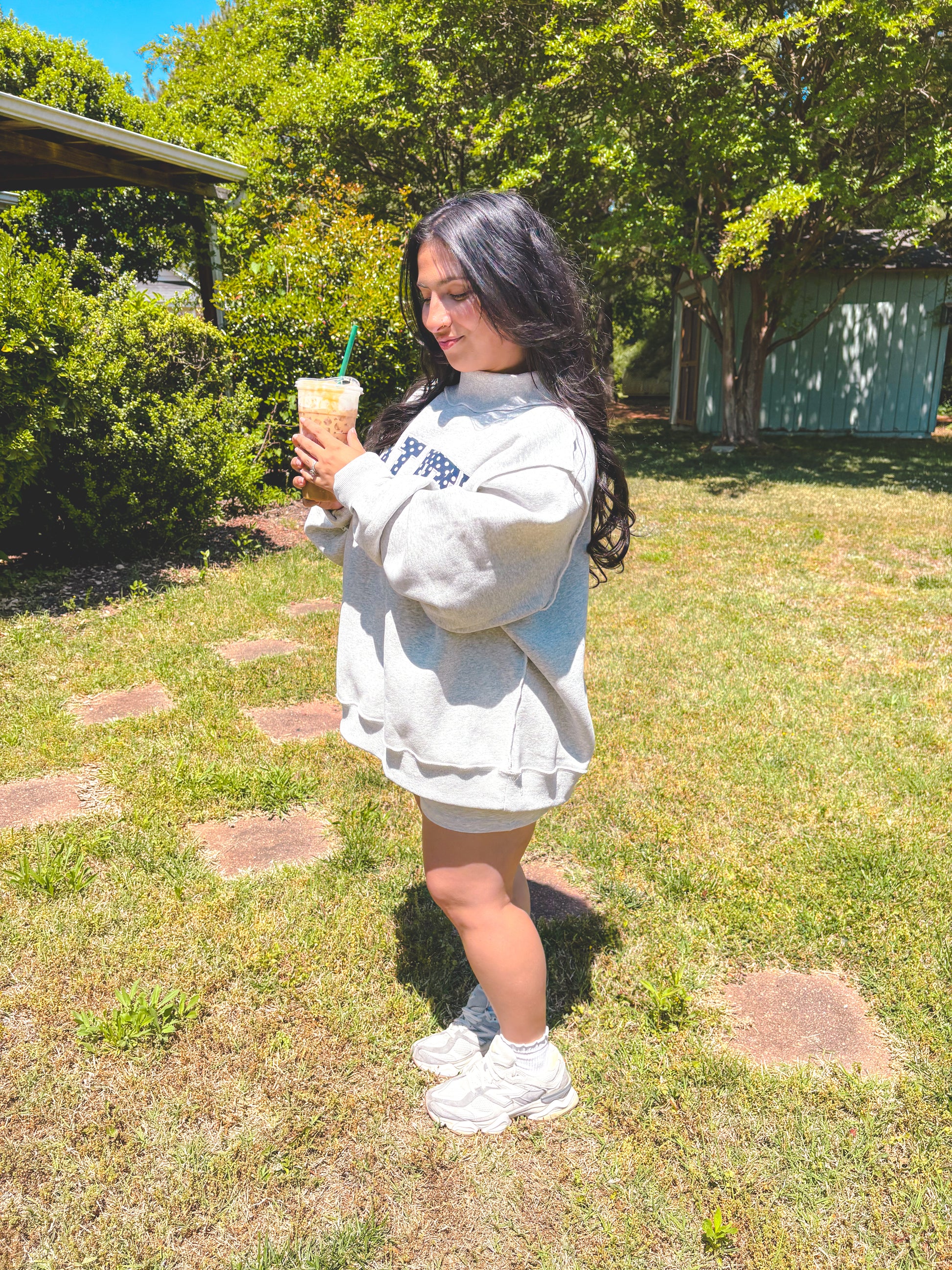 Person wearing a light gray sweatshirt with text, holding a drink outdoors.