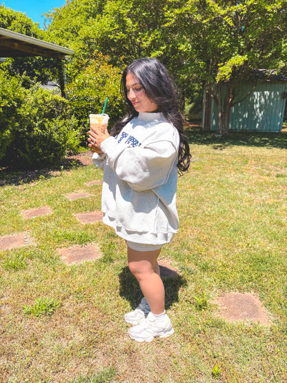 Person wearing a light gray sweatshirt with text, holding a drink outdoors.