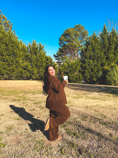 Person in a brown pants set outfit taking a picture in a park with trees and clear blue sky.