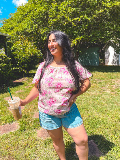 Woman in a floral shirt and blue shorts holding a drink outdoors.