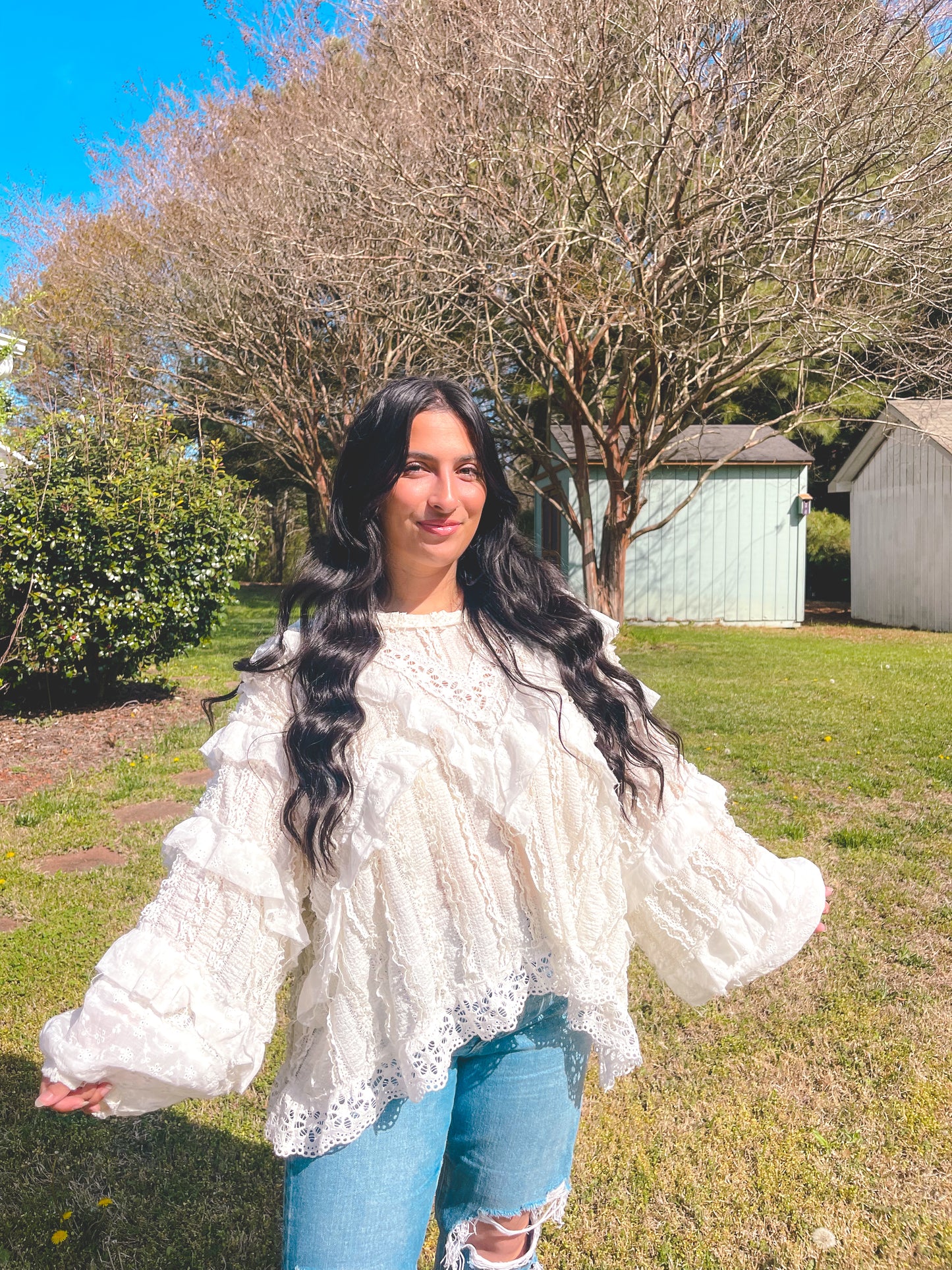 Woman wearing a white lace top and blue jeans standing outdoors with trees and a clear sky in the background.