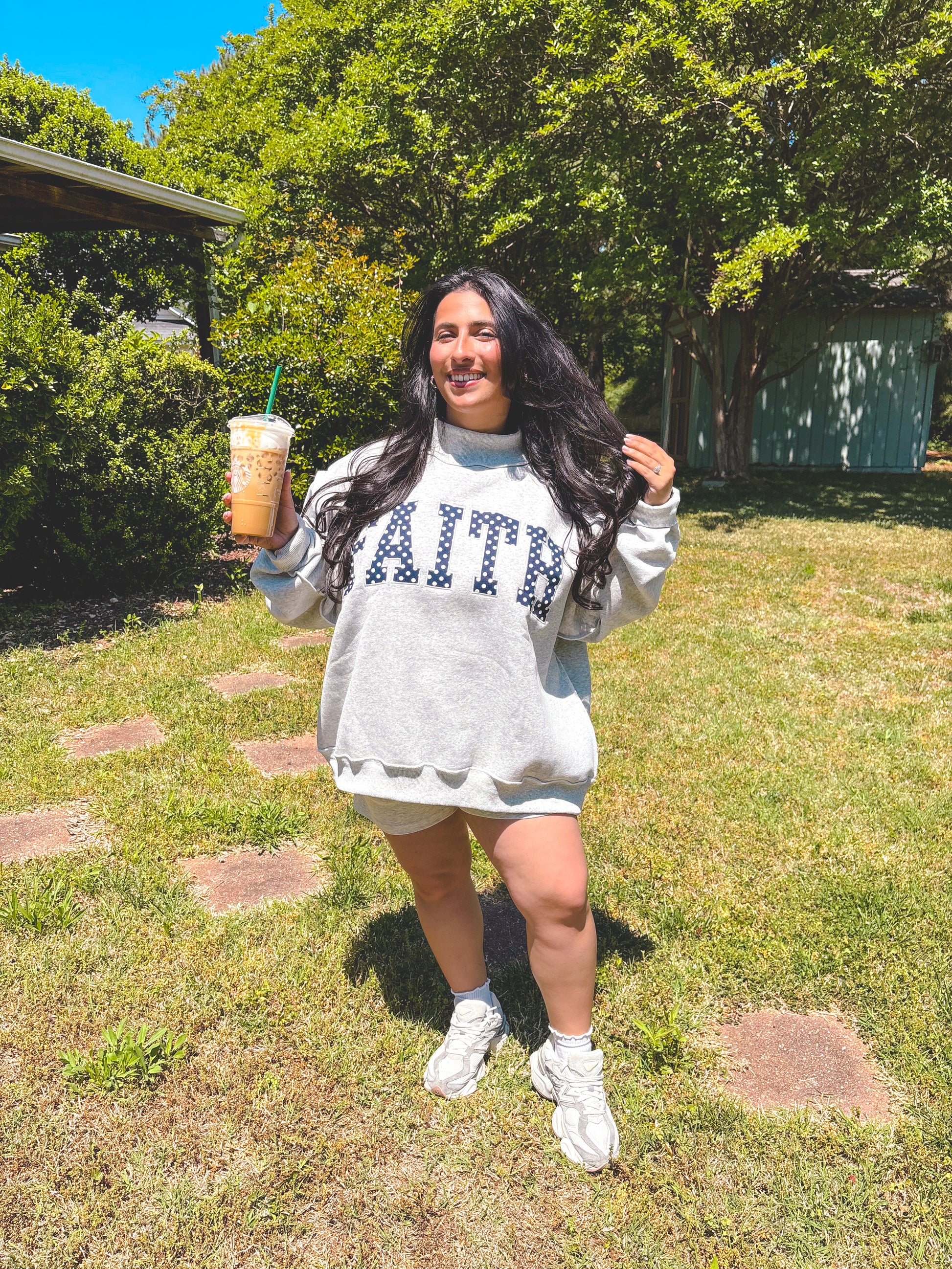 Woman in a gray sweatshirt with 'FAITH' holding an iced drink outdoors.