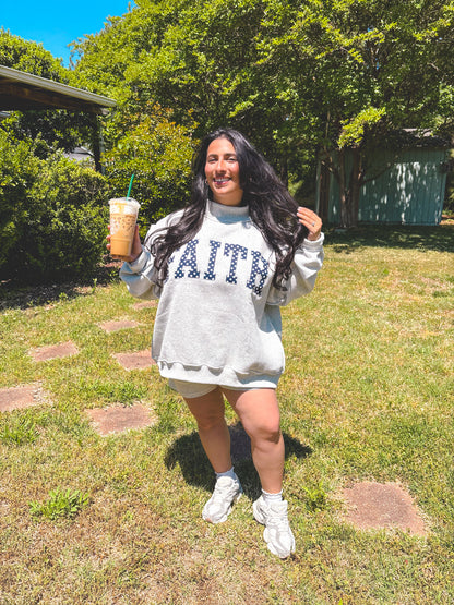 Woman in a gray sweatshirt with 'FAITH' holding an iced drink outdoors.