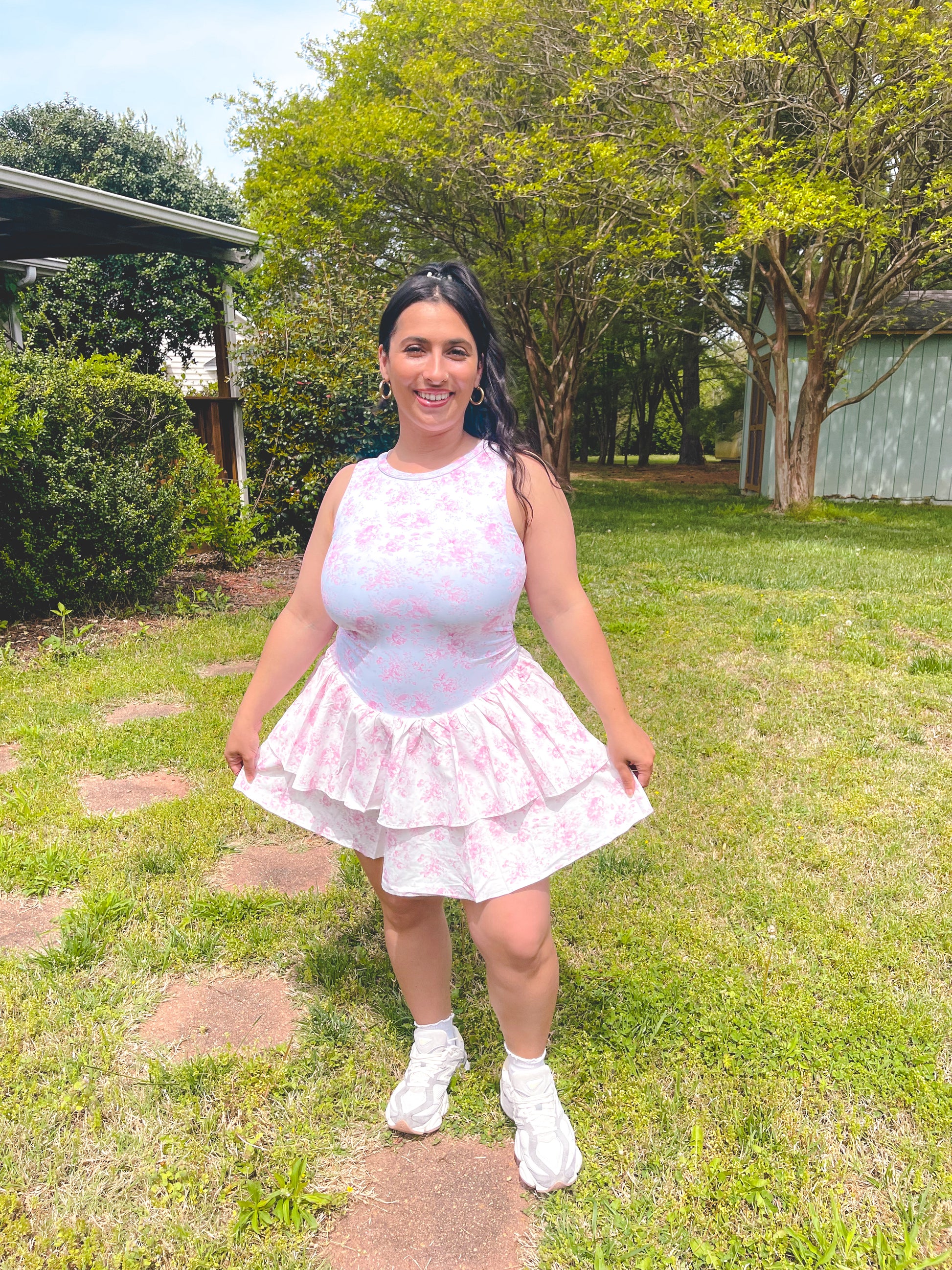 Woman in a light pink top and skirt standing in a grassy area with trees in the background