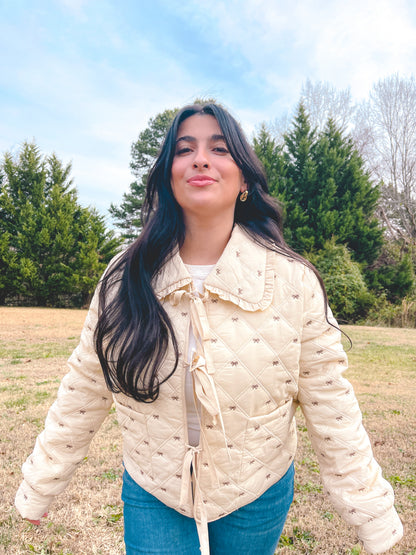 Woman wearing a cream jacket that has small brown bows all over and it is paired with blue jeans standing in a field with trees in the background