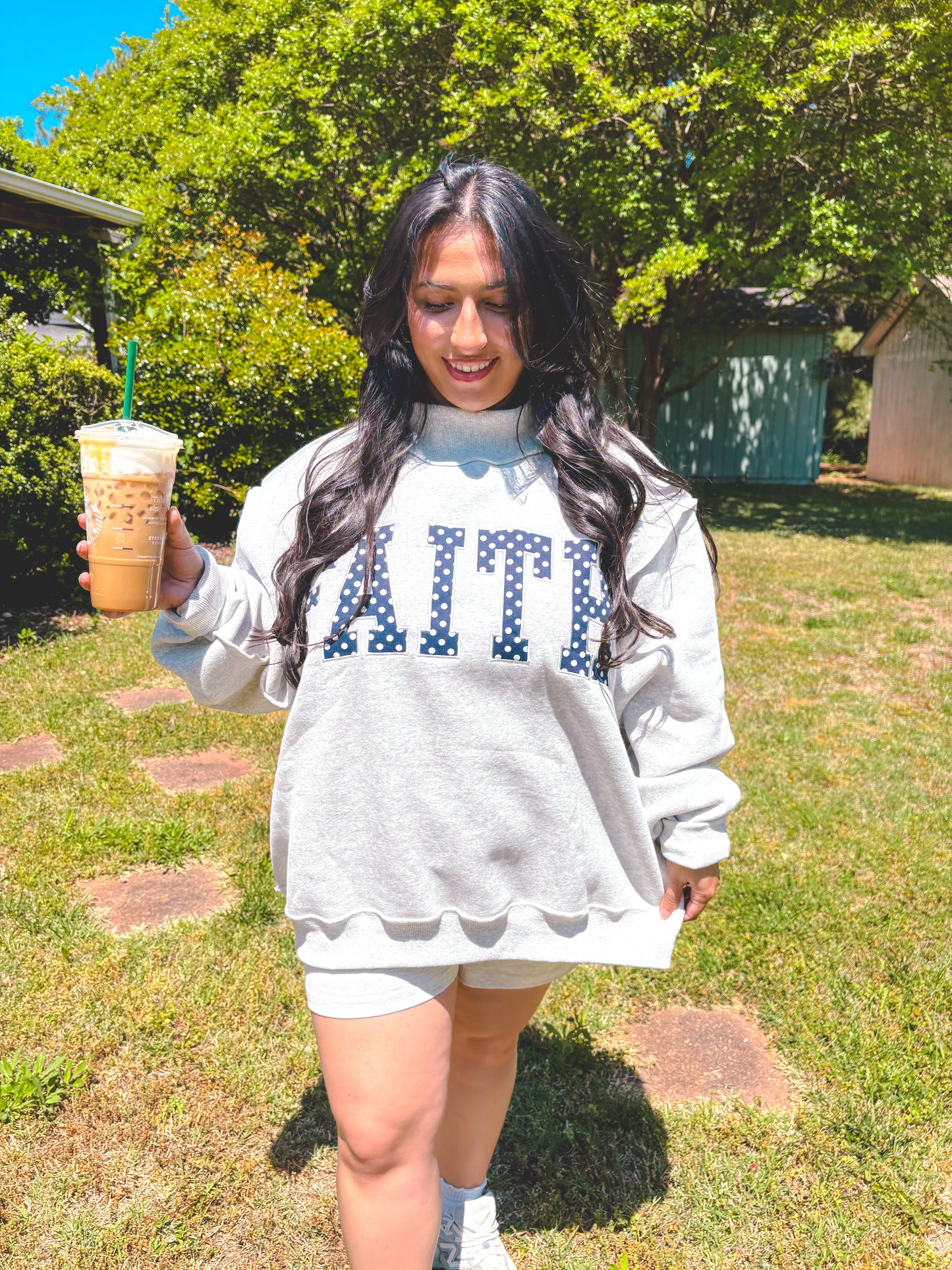 Woman wearing a sweatshirt with 'FAITH' printed on it, holding a drink outdoors.