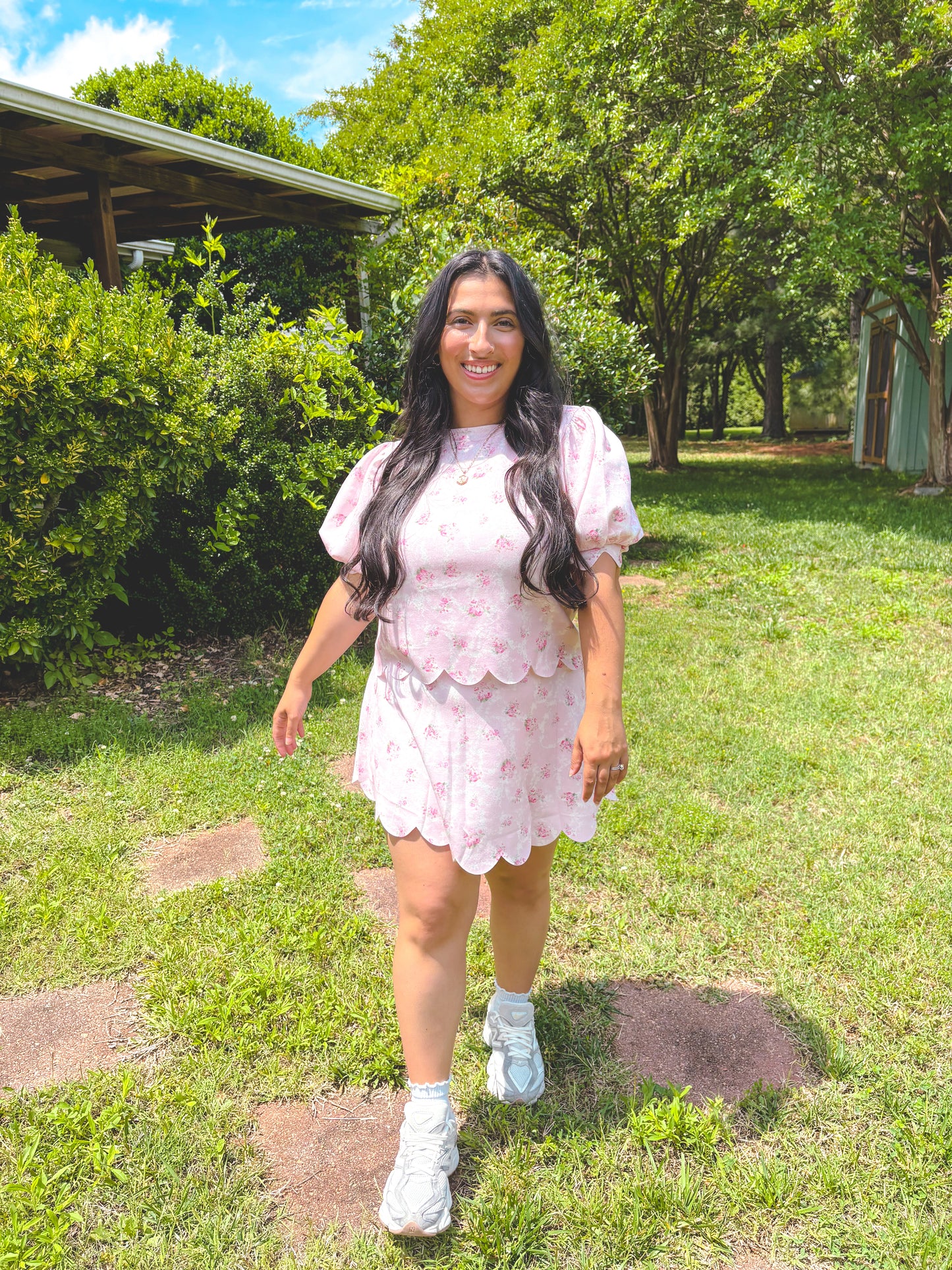 Woman in a pink dress standing in a grassy area with trees and a building in the background.