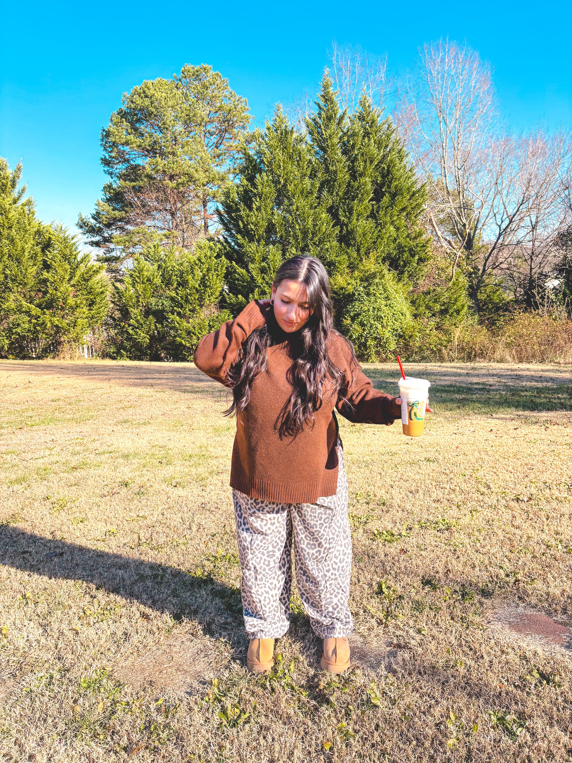Model in a brown sweater and leopard print pants holding a drink outdoors, showcasing fall fashion style.