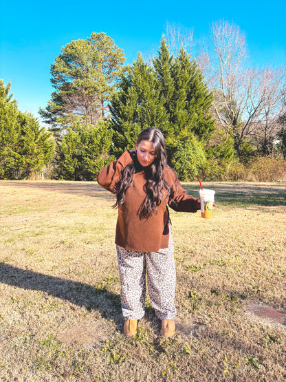 Model in a brown sweater and leopard print pants holding a drink outdoors, showcasing fall fashion style.