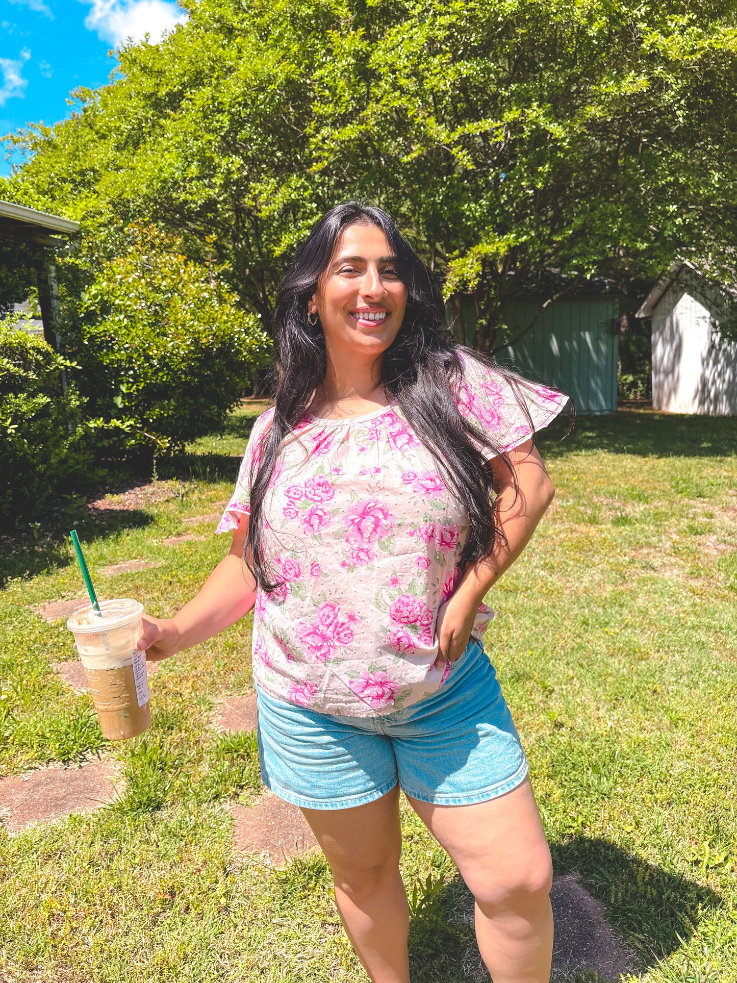 Woman in a floral shirt and shorts holding a drink in a grassy area with trees.