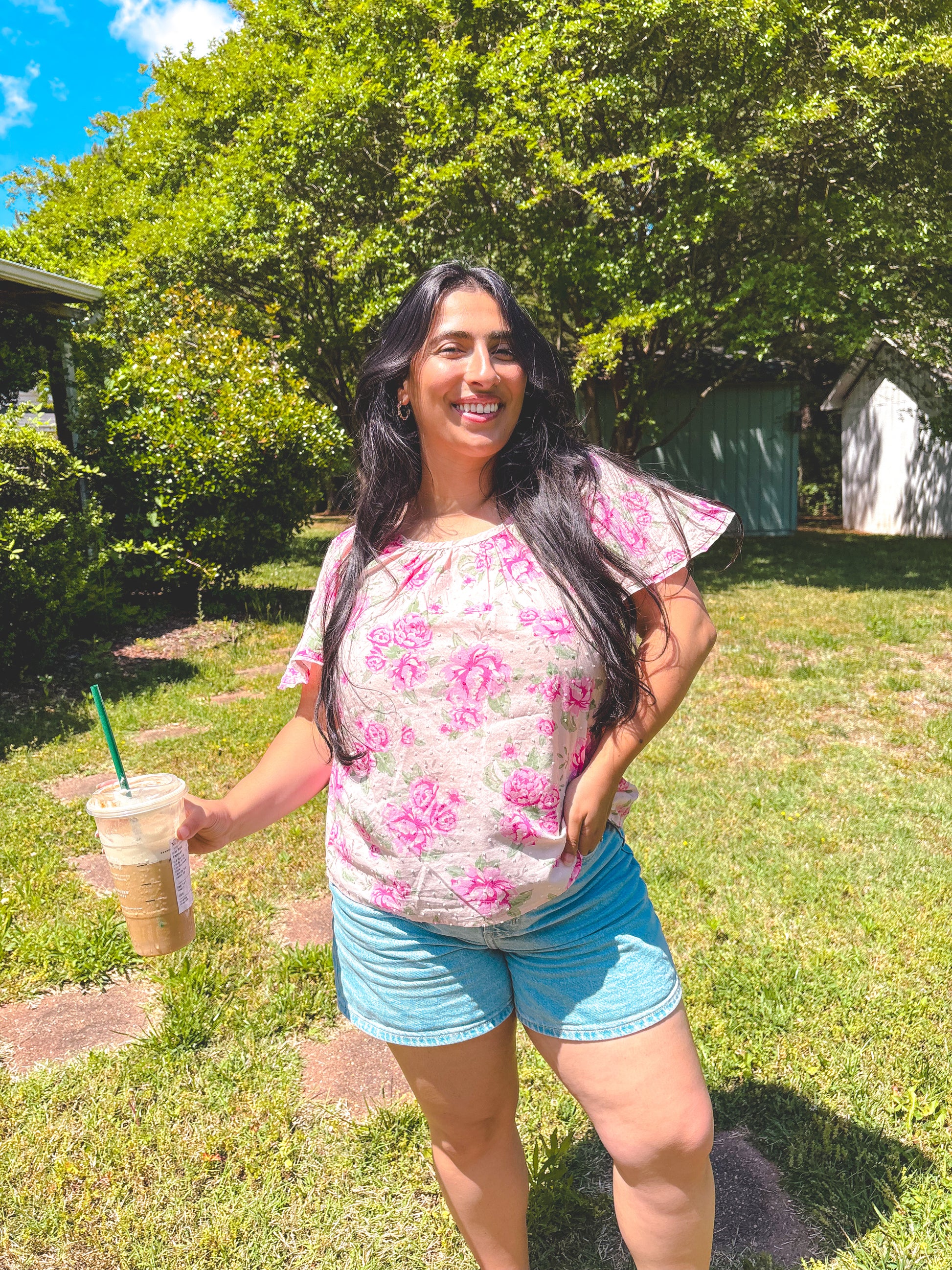 Woman in a floral shirt and shorts holding a drink in a grassy area with trees.