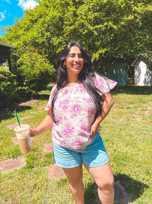 Woman in a floral shirt and shorts holding a drink in a grassy area with trees.