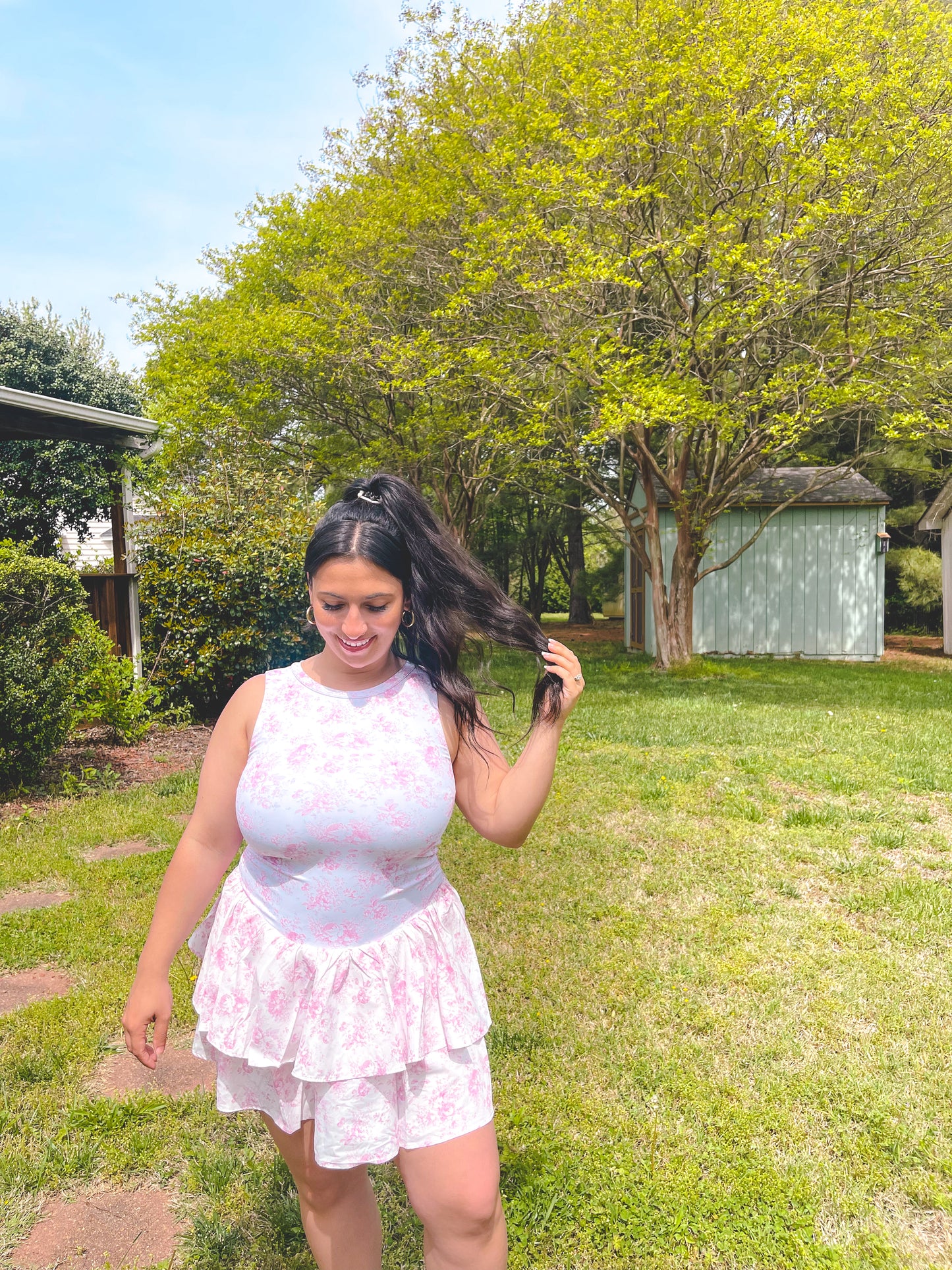 Woman in a pink top and white skirt standing in a grassy area with trees in the background
