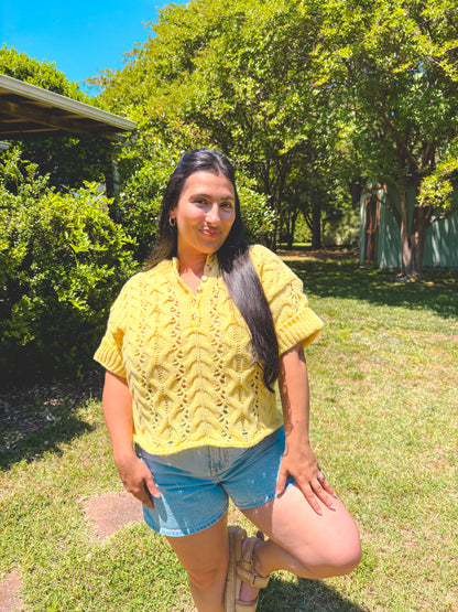Woman in a yellow top and denim shorts standing in a park with trees and grass.