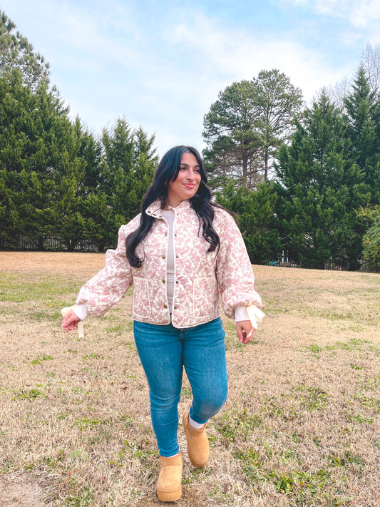 Woman in a floral jacket paired with blue jeans walking in a field with trees in the background