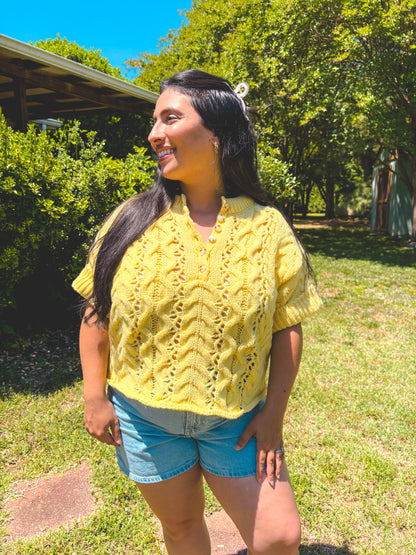 Woman wearing a yellow knitted shirt and denim shorts standing outdoors on a sunny day.
