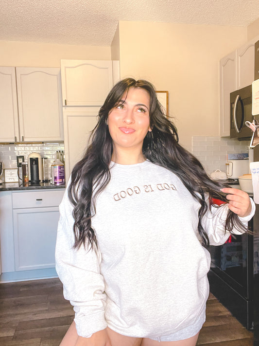 Woman in a kitchen wearing a grey sweatshirt with text saying God is good, holding her hair.