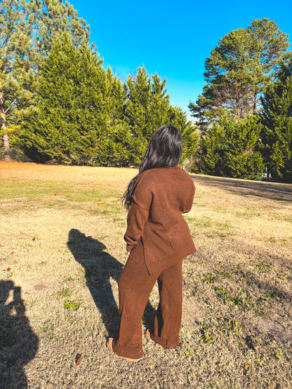 Person wearing a brown outfit pant set standing in a park with trees and clear blue sky.