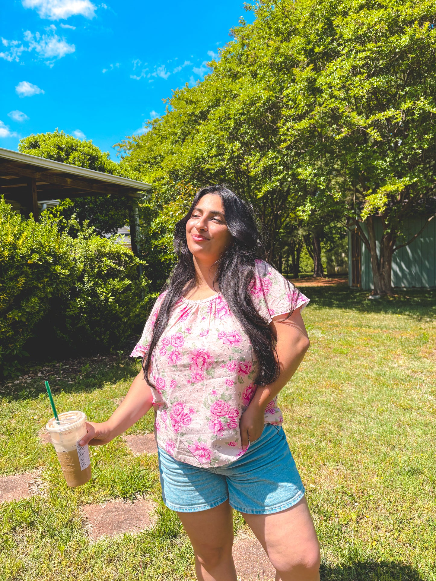 Woman in a floral shirt and shorts holding a drink outdoors on a sunny day