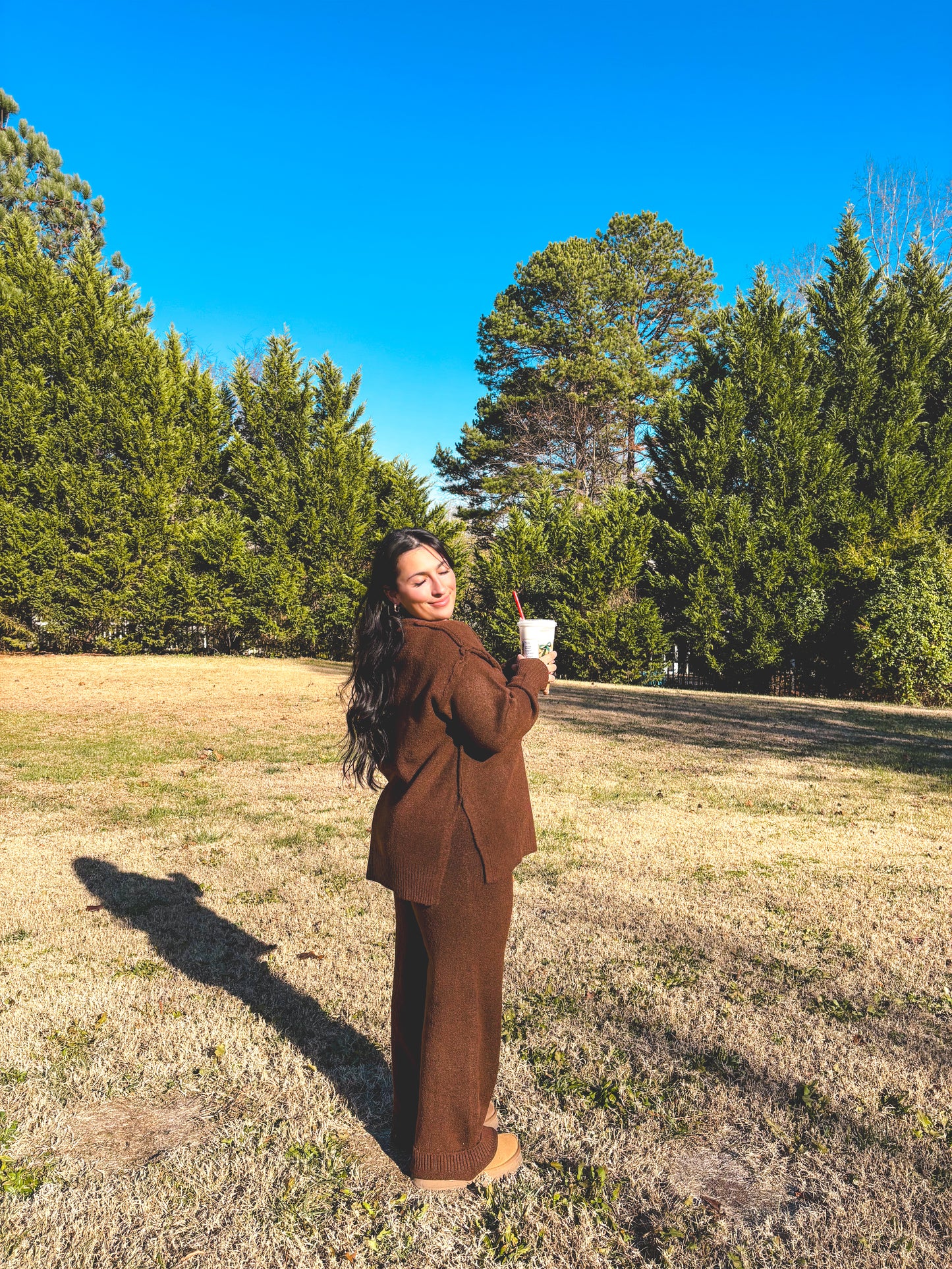 Woman in a brown outfit pant set standing in a field with trees and blue sky