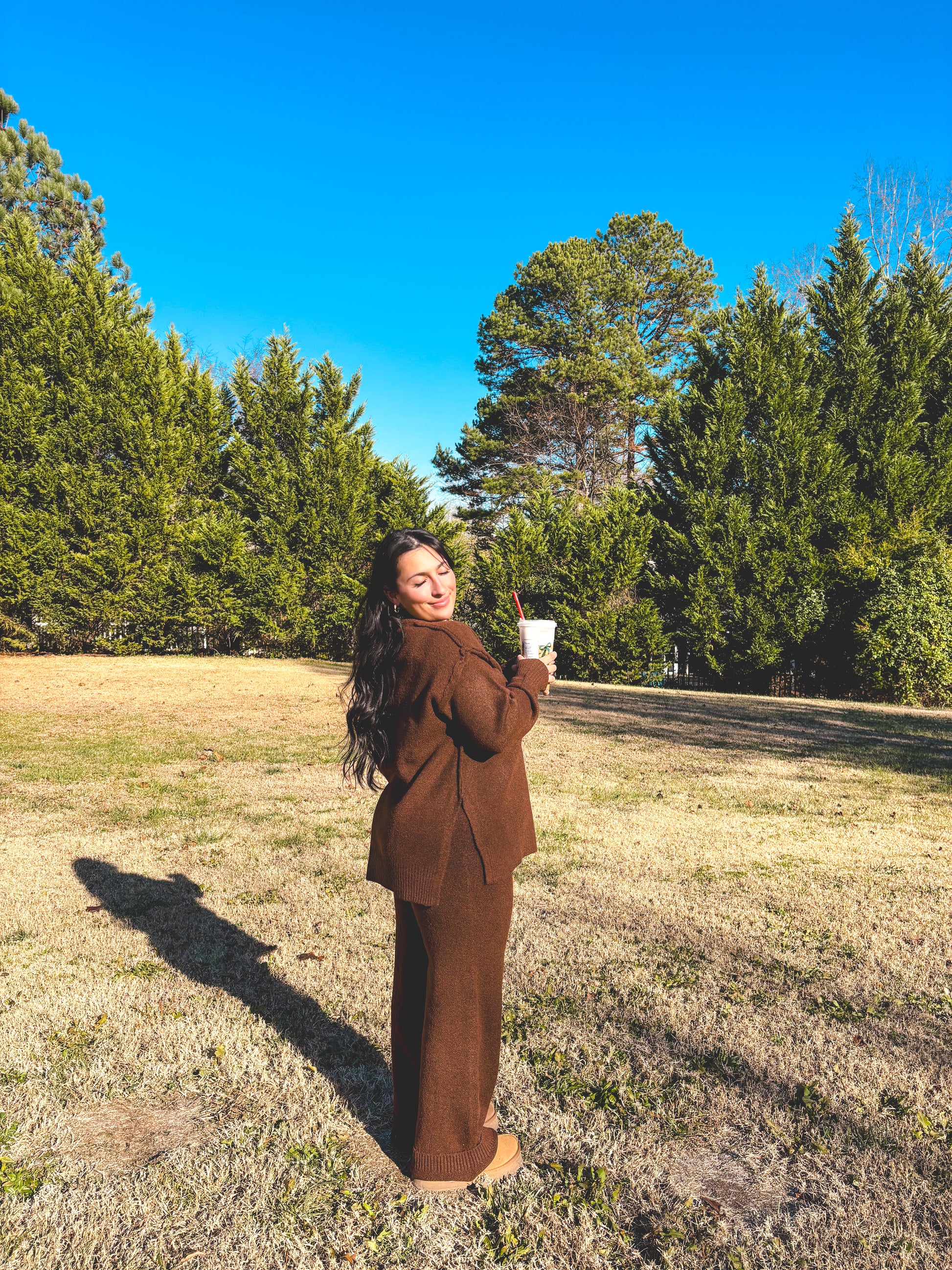 Woman in a brown outfit pant set standing in a field with trees and blue sky