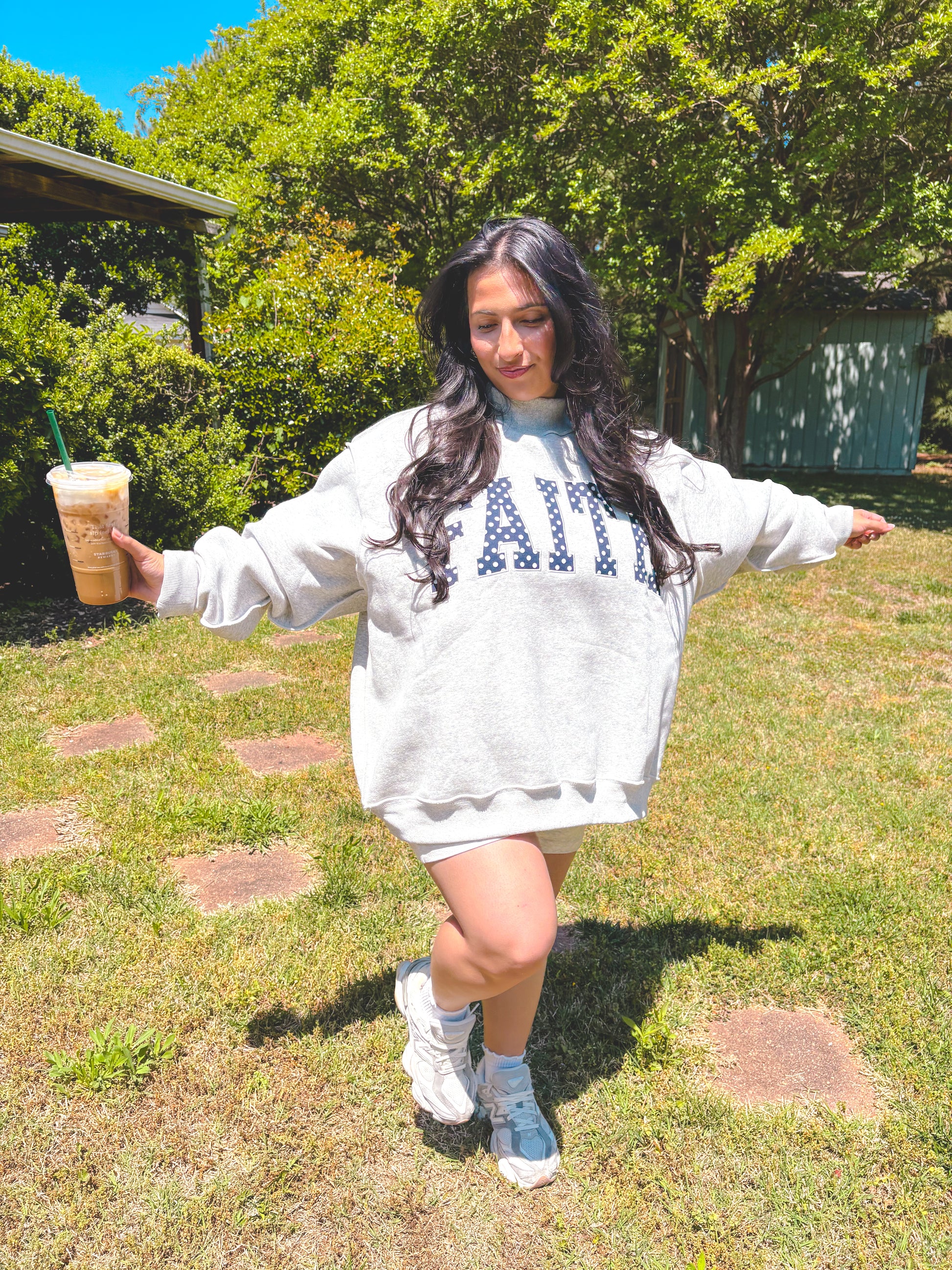 Person wearing a white sweatshirt with text, holding a drink outdoors.