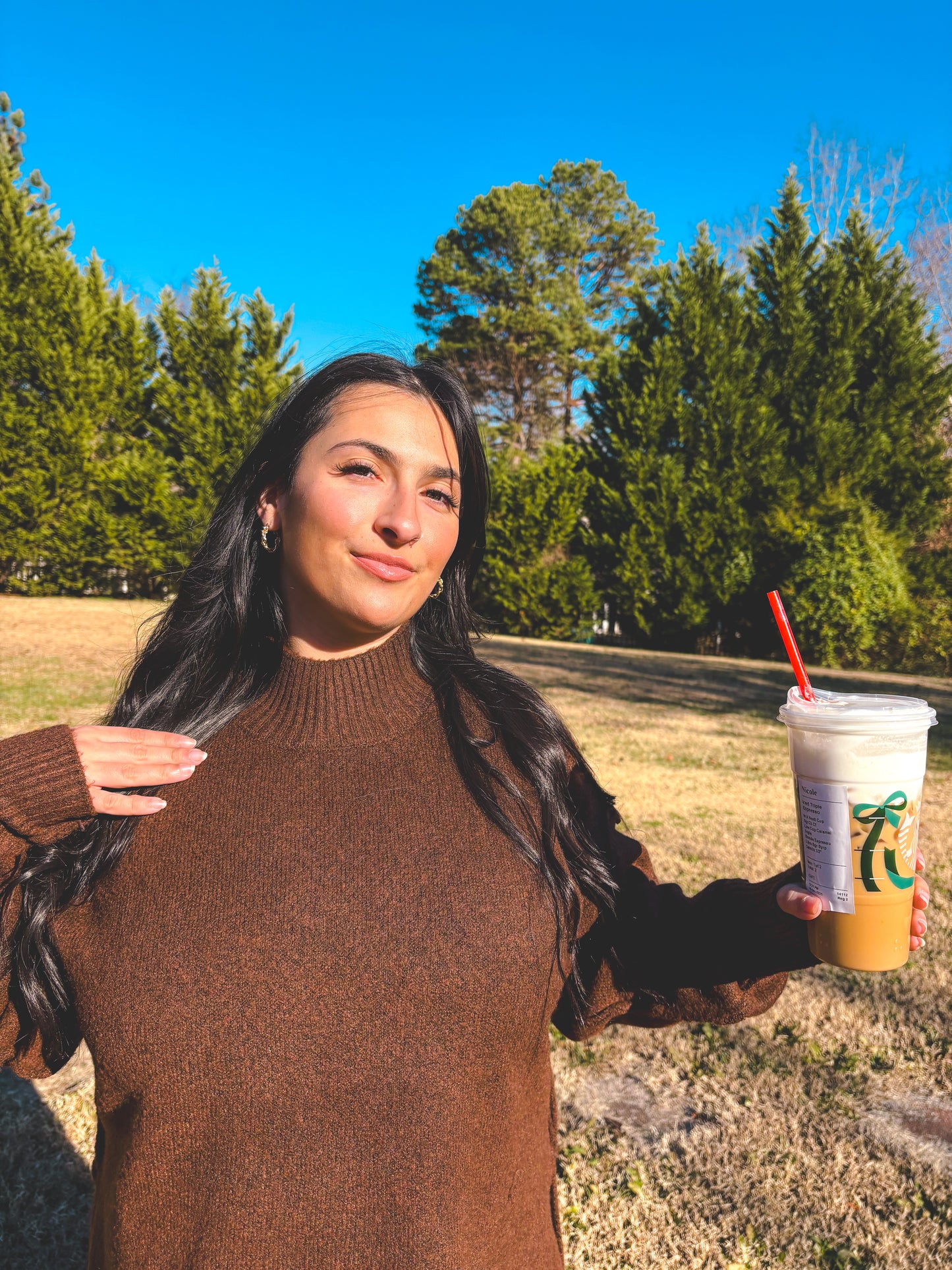Woman in a brown sweater holding a drink with a straw outdoors on a sunny day