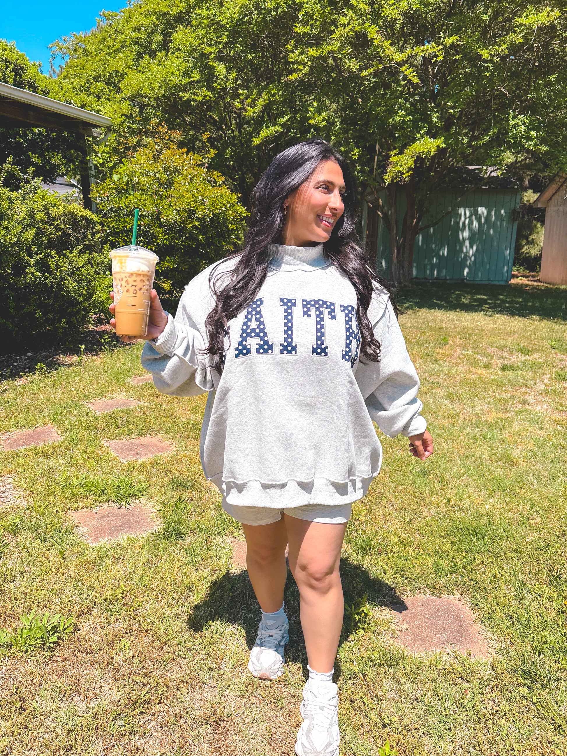 Woman in a gray sweatshirt with 'FAITH' printed on it, holding a drink outdoors.