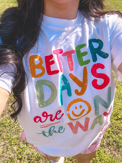 Person wearing a shirt with 'Better Days are on the Way' text in colorful letters outdoors.