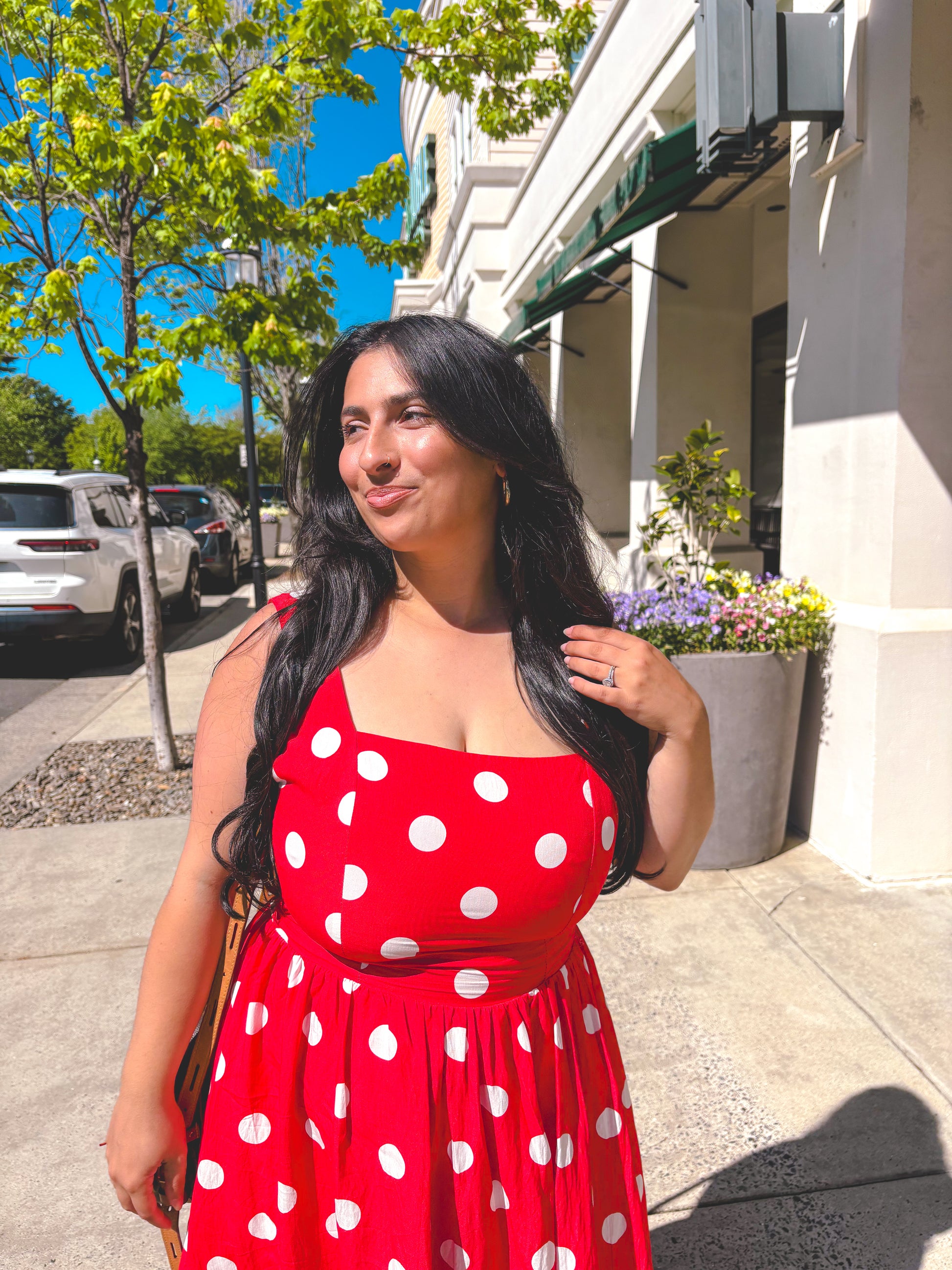 Woman in a red polka dot dress standing on a sidewalk with trees and buildings in the background.