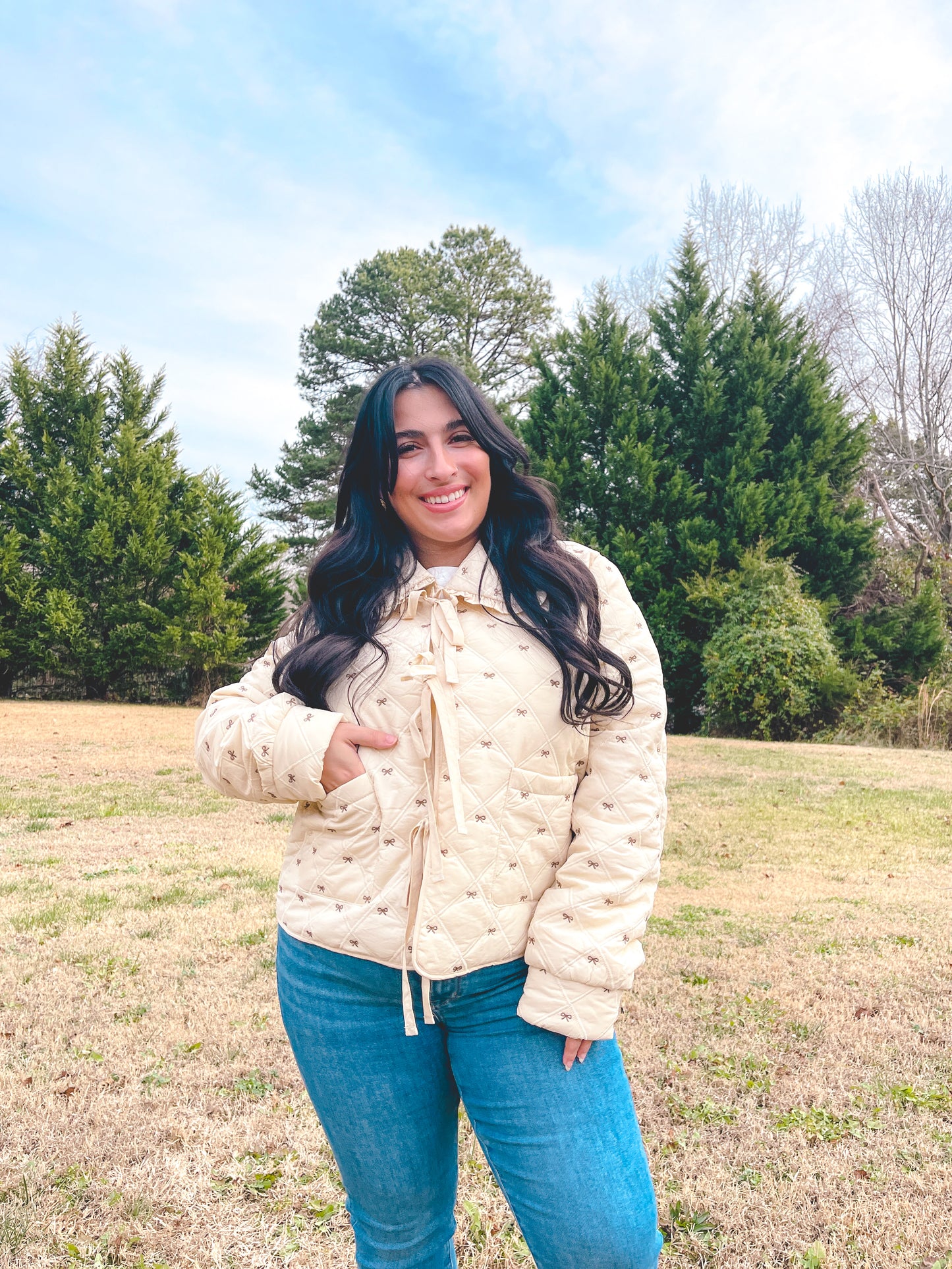 Woman wearing a cream jacket that has small brown bows all over and it is paired with blue jeans standing in a field with trees in the background