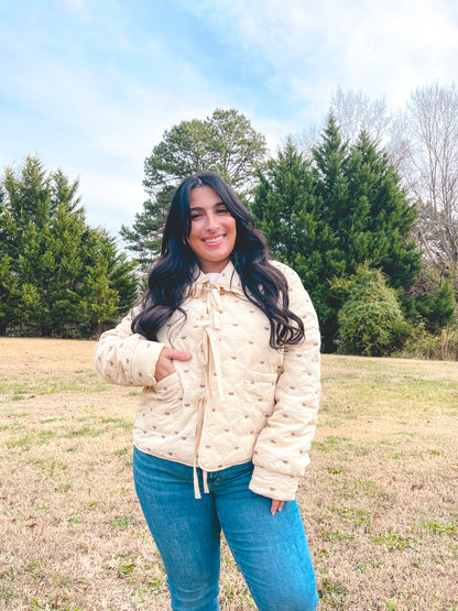 Woman wearing a cream jacket that has small brown bows all over and it is paired with blue jeans standing in a field with trees in the background