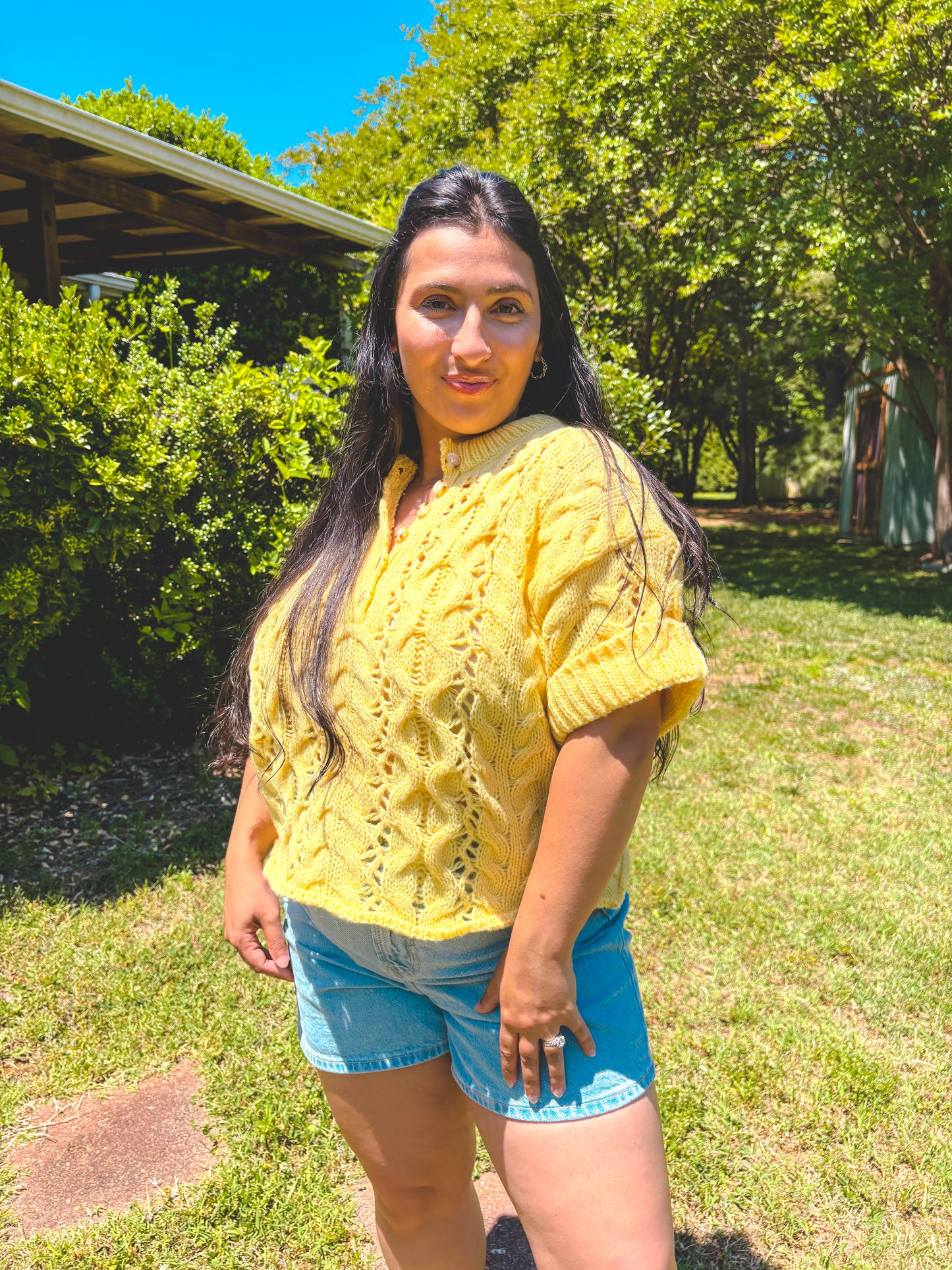 Woman wearing a yellow knitted sweater and blue shorts standing outdoors with greenery in the background