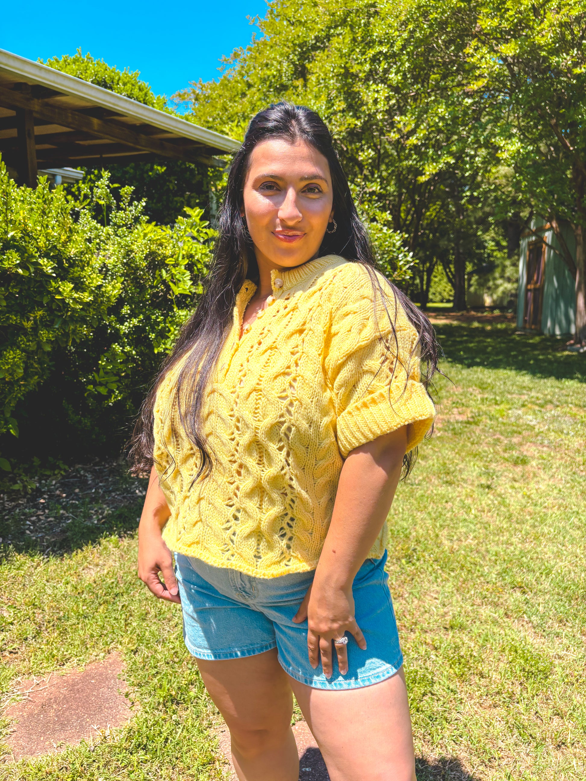 Woman wearing a yellow knitted sweater and blue shorts standing outdoors with greenery in the background