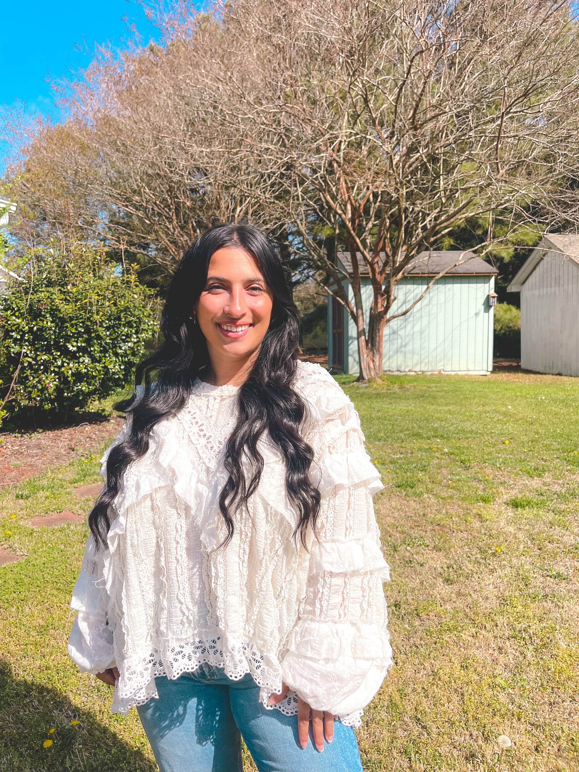 Woman in a white blouse standing outdoors with trees and a shed in the background