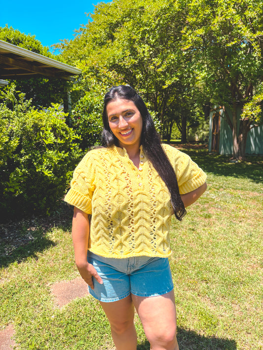 Woman wearing a yellow shirt and denim shorts standing in a grassy area with trees.