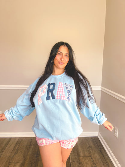 Woman wearing a light blue sweatshirt with 'PRAY' text in a room with gray walls and wooden floor.