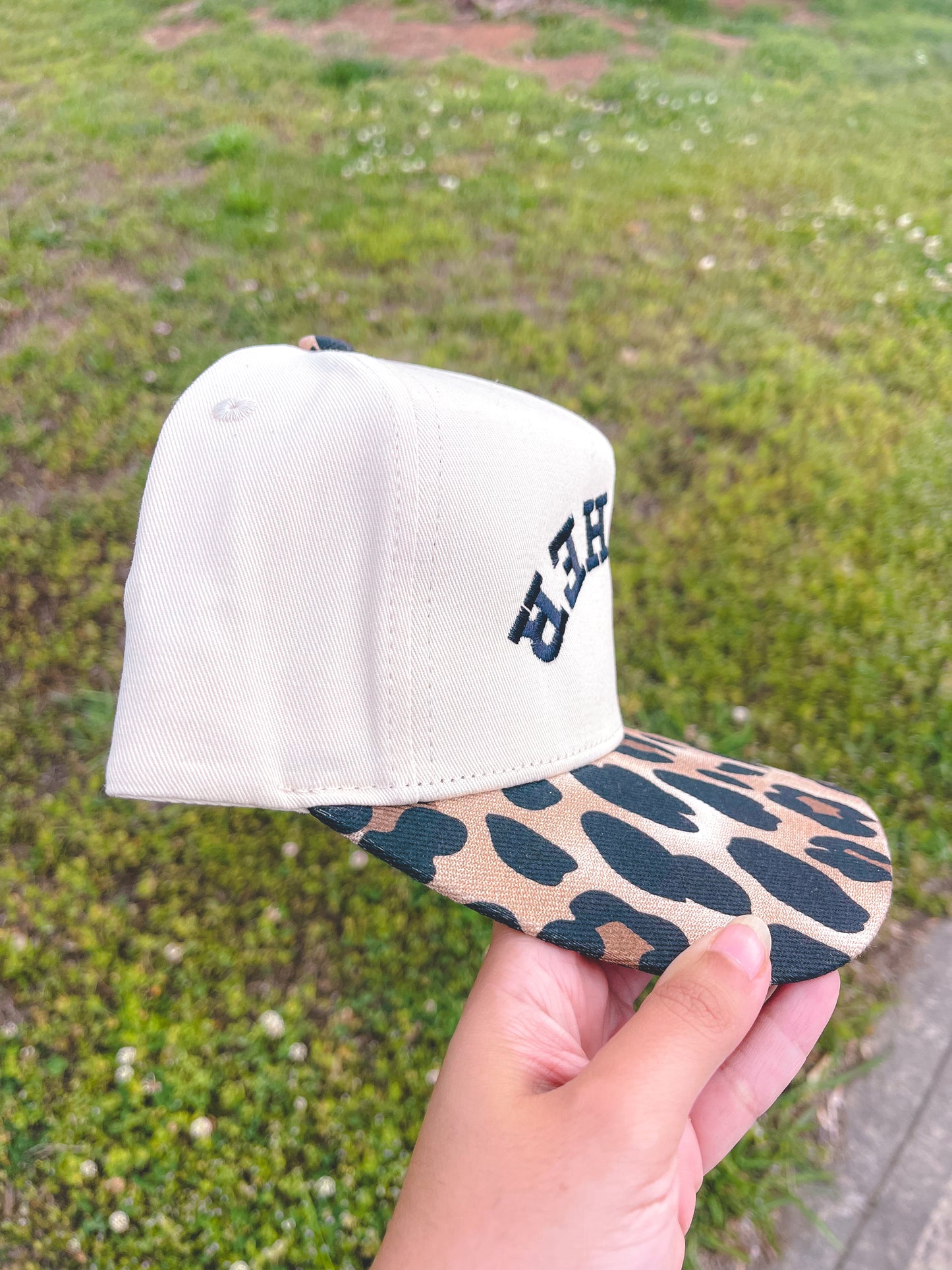 White cap with leopard print brim held in front of a grassy background