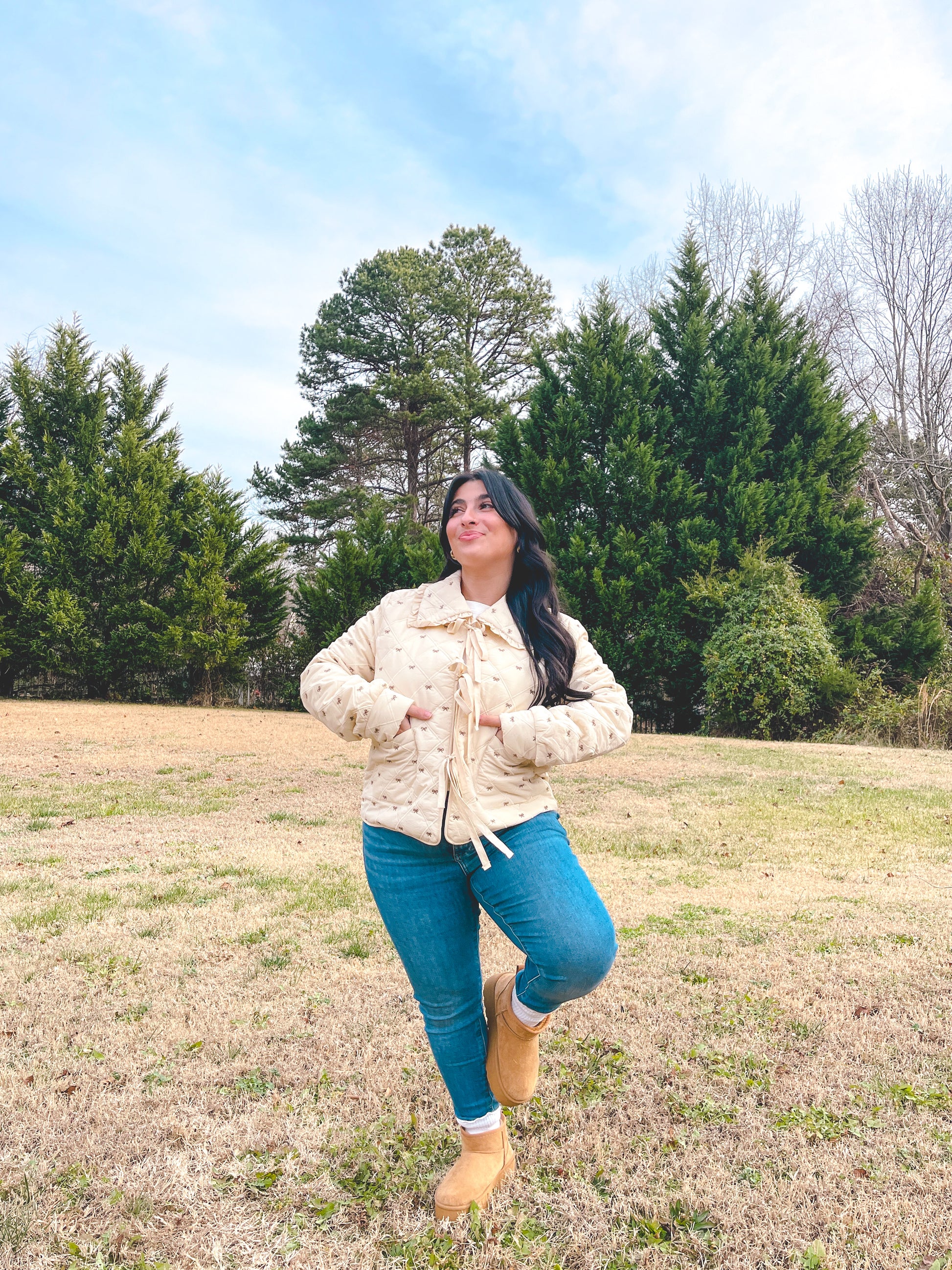 Woman in a beige jacket and blue jeans standing in a field with trees in the background