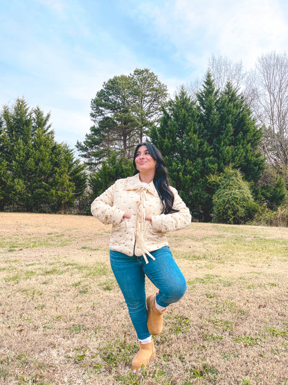 Woman in a beige jacket and blue jeans standing in a field with trees in the background