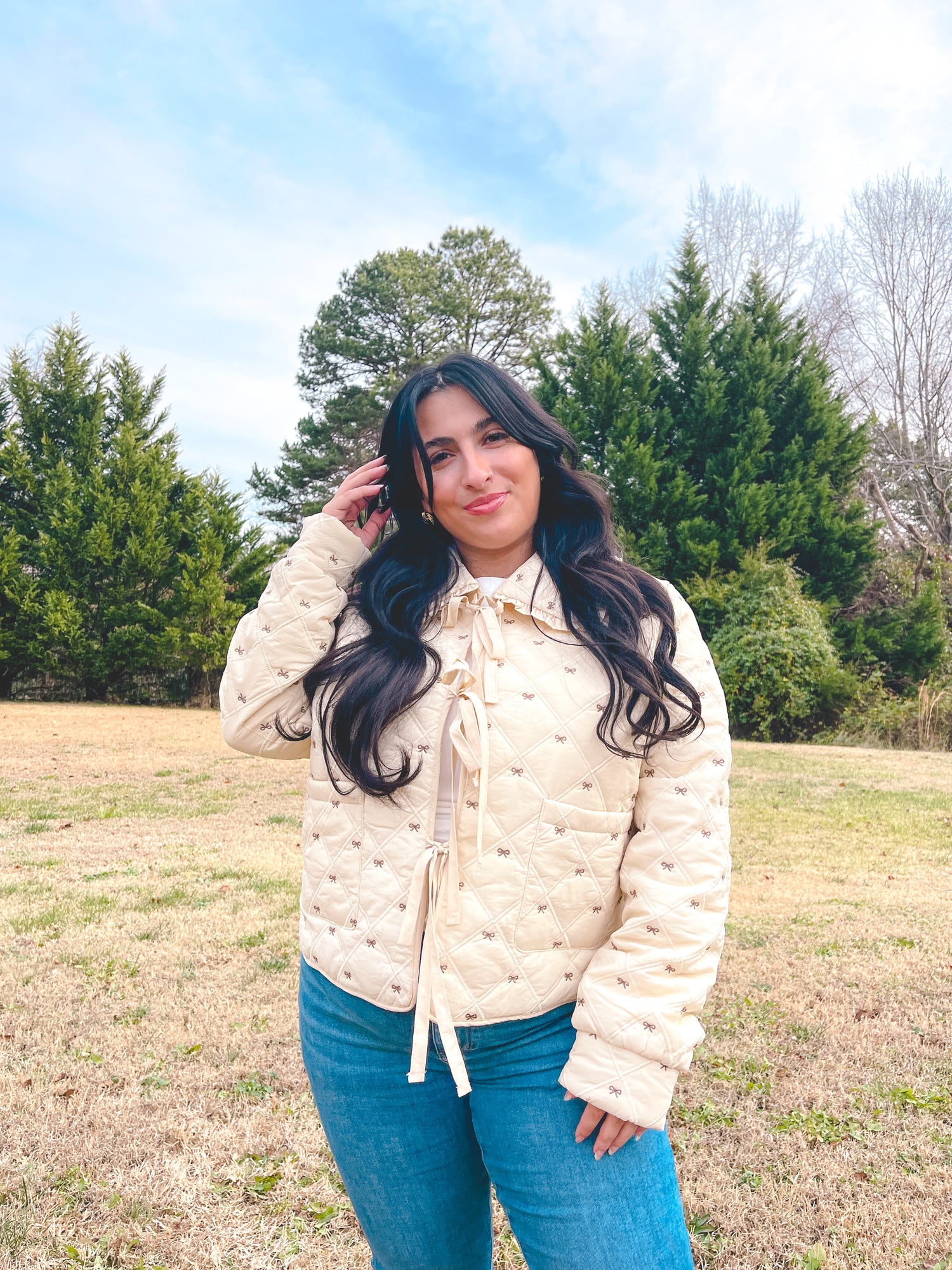 Woman wearing a cream jacket that has small brown bows all over and it is paired with blue jeans standing in a field with trees in the background