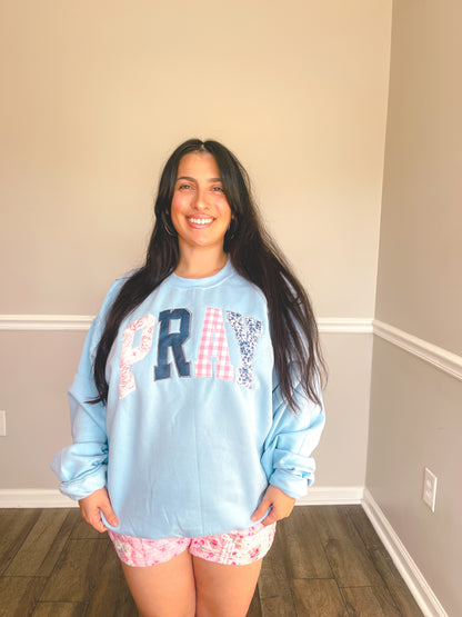 Woman wearing a light blue sweatshirt with 'PRAY' printed on it, standing in a room with a neutral wall.