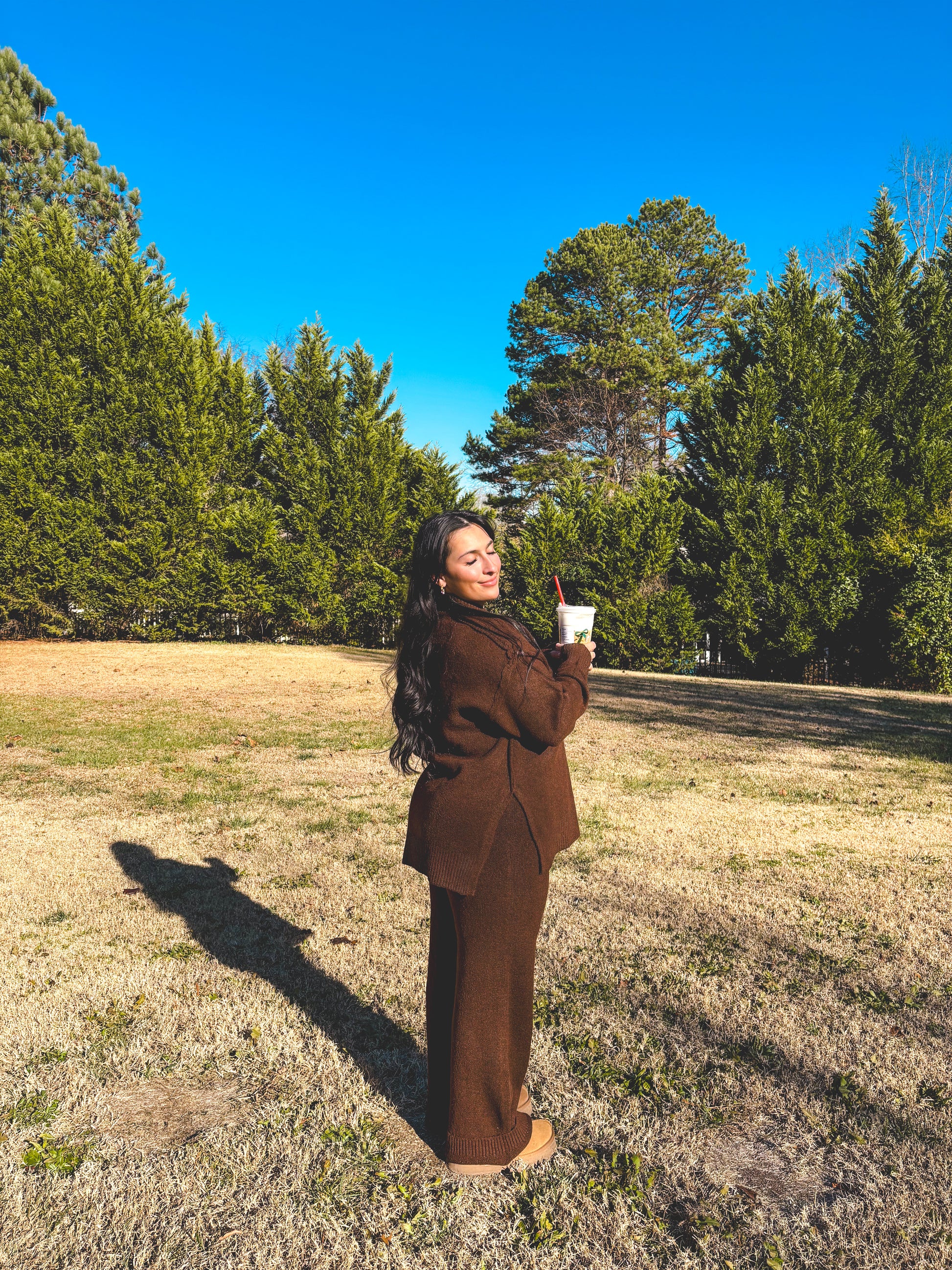 Woman in a brown pants set outfit standing in a field with trees and blue sky.