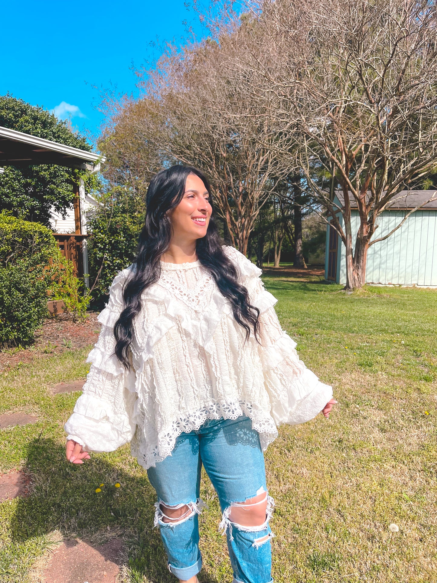 Woman wearing a white lace top and ripped jeans standing outdoors on a sunny day.