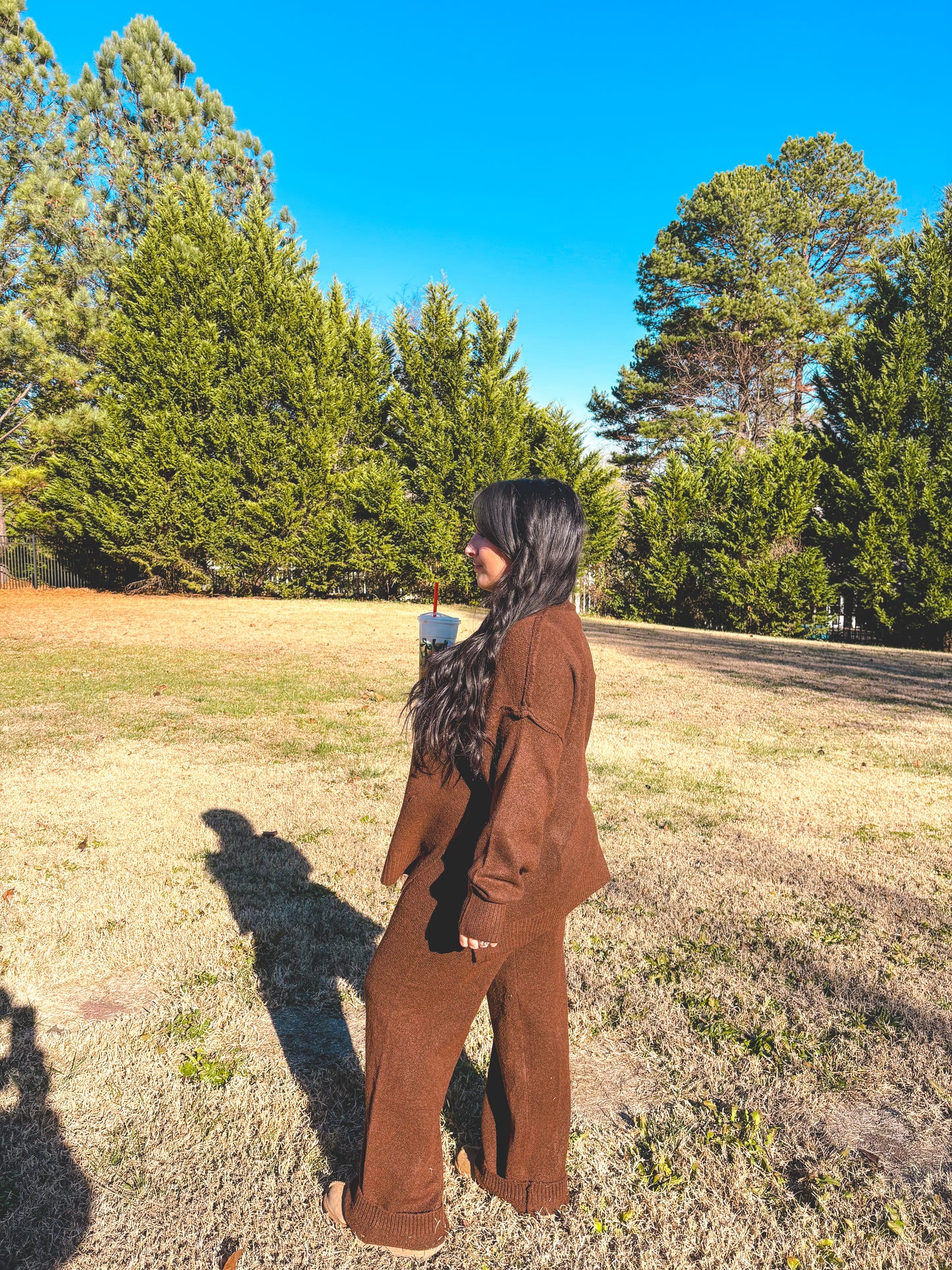 Person wearing a brown pant outfit set standing in a field with trees in the background