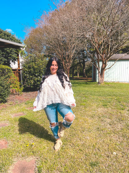 Woman in a white lace top and ripped jeans walking on grass with trees and a house in the background.